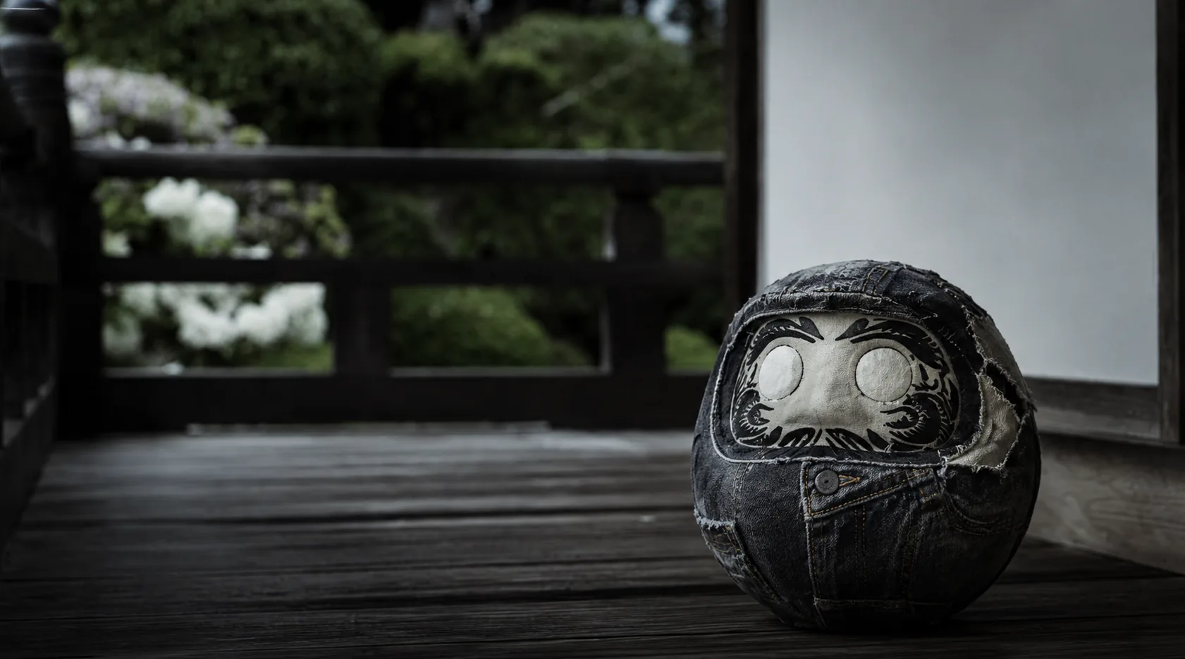 日本家屋の縁側に佇む、ブラックデニムを縫合したダルマ彫像。背景に庭の緑と障子。